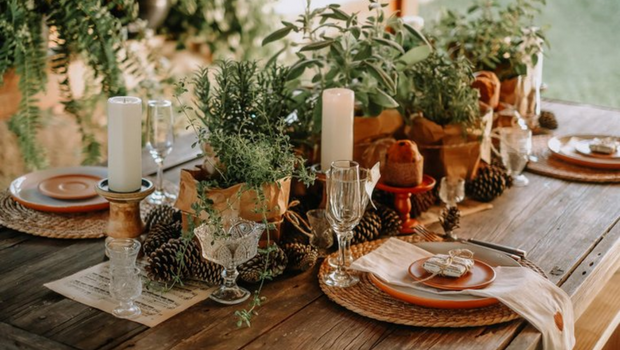 Mesa de madeira com pratos redondos. Ao centro castiçais de cristal com velas e panetones, arranjo de pinhas com alecrim.