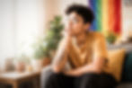 LGBTQ+ adult sitting thoughtfully on a couch in a softly lit room with pride flag colors in the background, appearing emotionally exhausted and reflective.