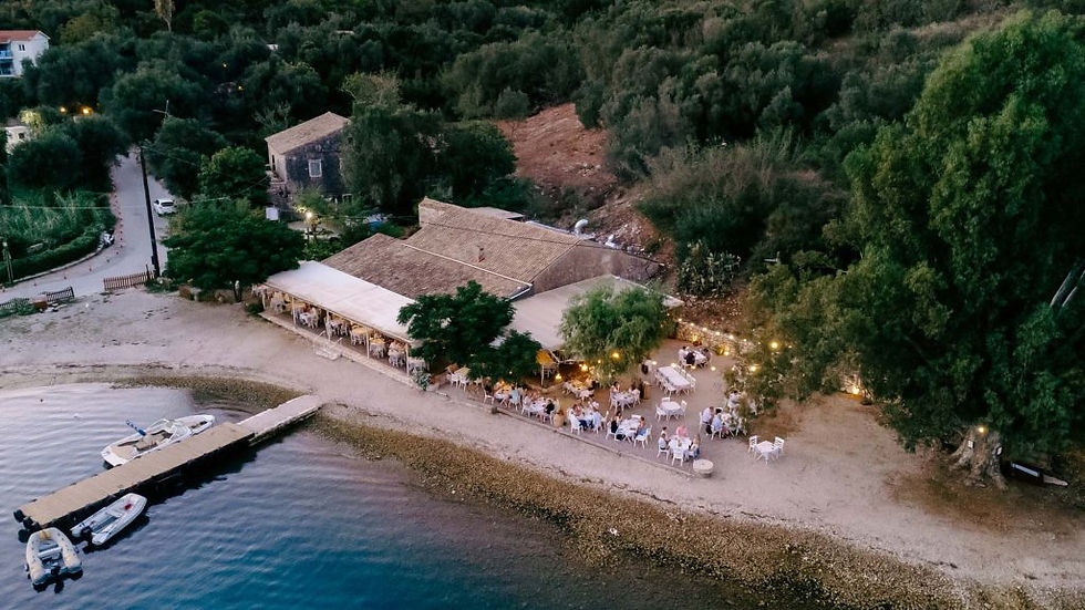 Wide landscape aerial photo of Eucalyptus seaside taverna on the beach in Corfu at dusk, with warm lights, trees, and shoreline.