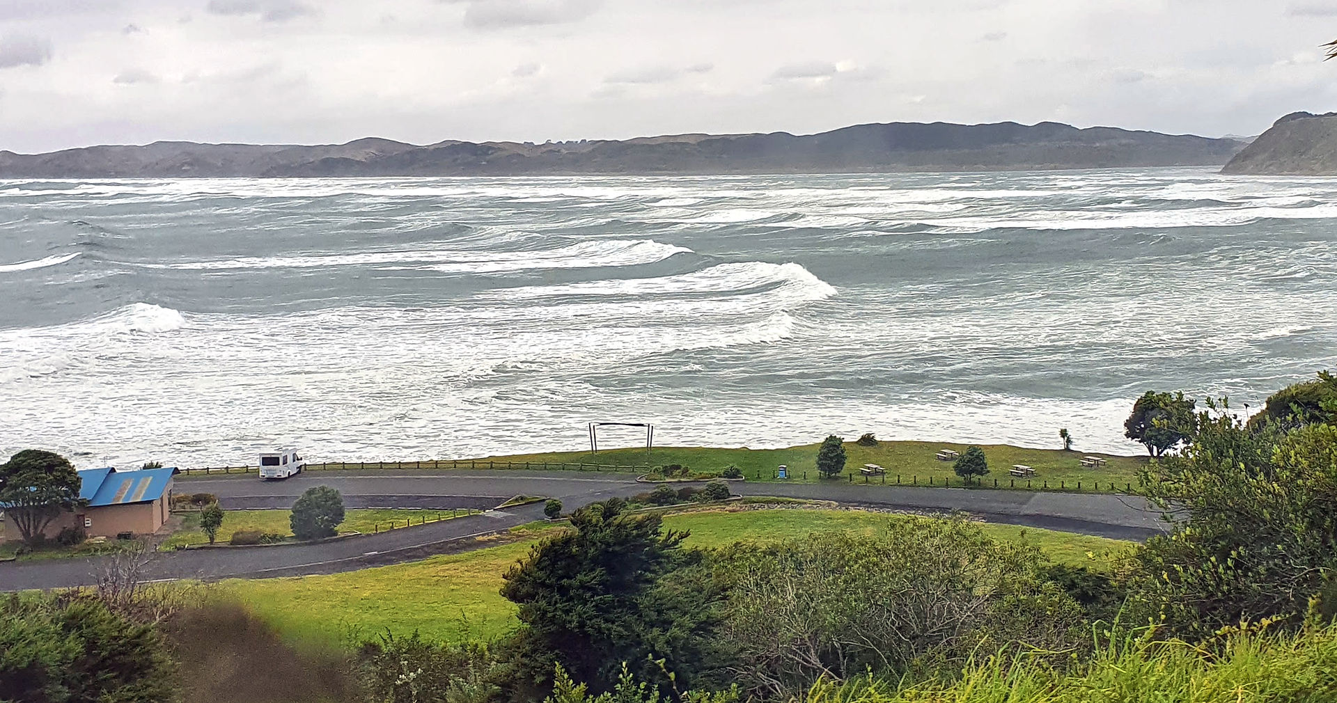 Choppy ocean waves, grassy coast with white structure, distant hills