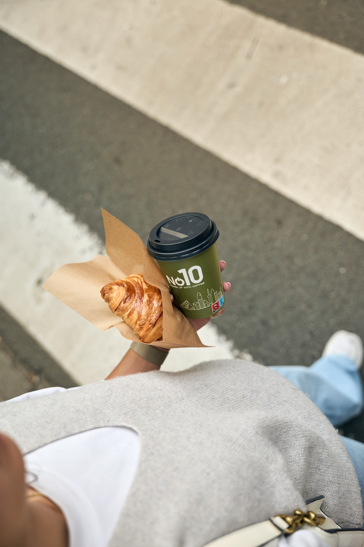 Person holding coffee cup and croissant on street.