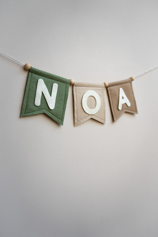 Thumbnail: Muur waar een wolvilten handgemaakte slinger hangt van vlaggen in de kleuren groen, beige en zand met de letters 'NOA'