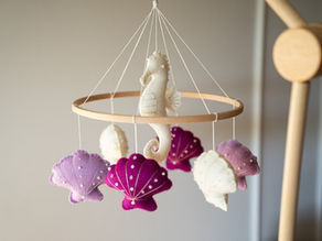 Babymobiel van hout met vilten figuren in de vorm van een zeepaardje en schelpen in paars, roze en crème tinten.