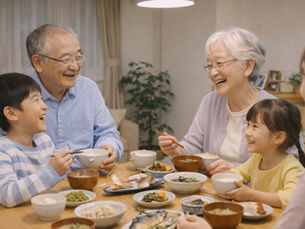 祖父母と孫たちが食卓を囲み、笑顔で会話を楽しみながら食事をしている様子。温かい家庭の雰囲気の中で、和食のおかずが並ぶ食卓を囲み、家族の団らんと食育の大切さが伝わる風景。