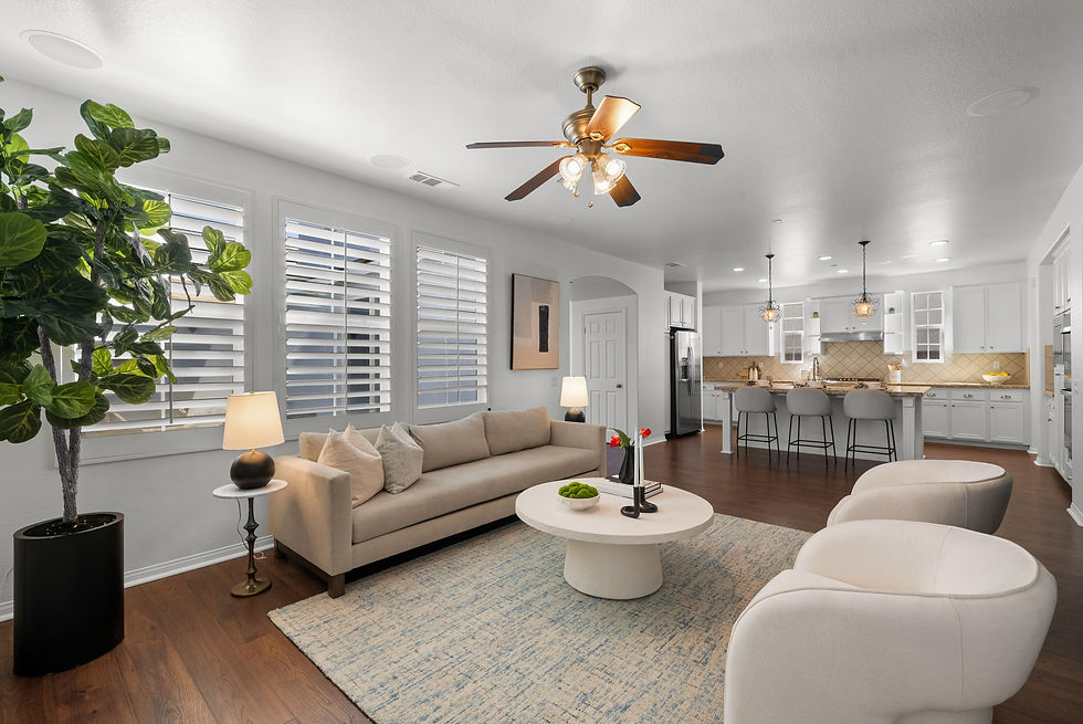 Modern living room with beige sofa, round white table, and two chairs. Ceiling fan above. Open kitchen with island and stools in background.