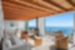 Cozy living room with ocean view, wood ceiling, white sofa, tan chairs, modern stove, and artwork. Large windows open to blue sea.