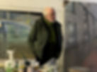 Man in green jacket and scarf with paintings behind him and art materials on the table in the foreground