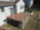 Bird's-eye view of a large deck construction in progress featuring brown composite decking material and white posts, adjacent to a white house with vinyl siding.