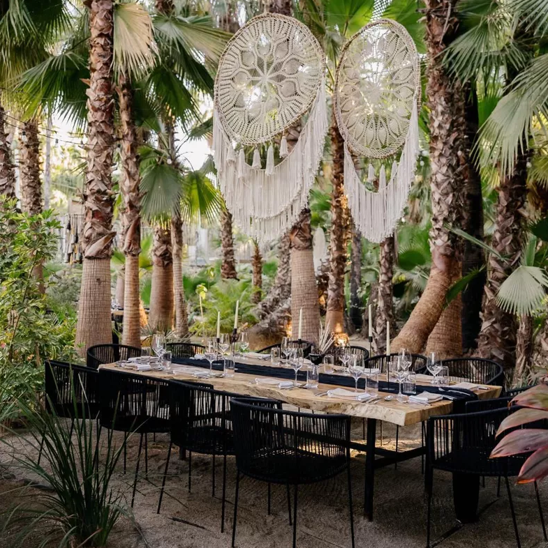 Tropical outdoor dining table with black chairs and dreamcatcher decor among palm trees, set for a stylish gathering