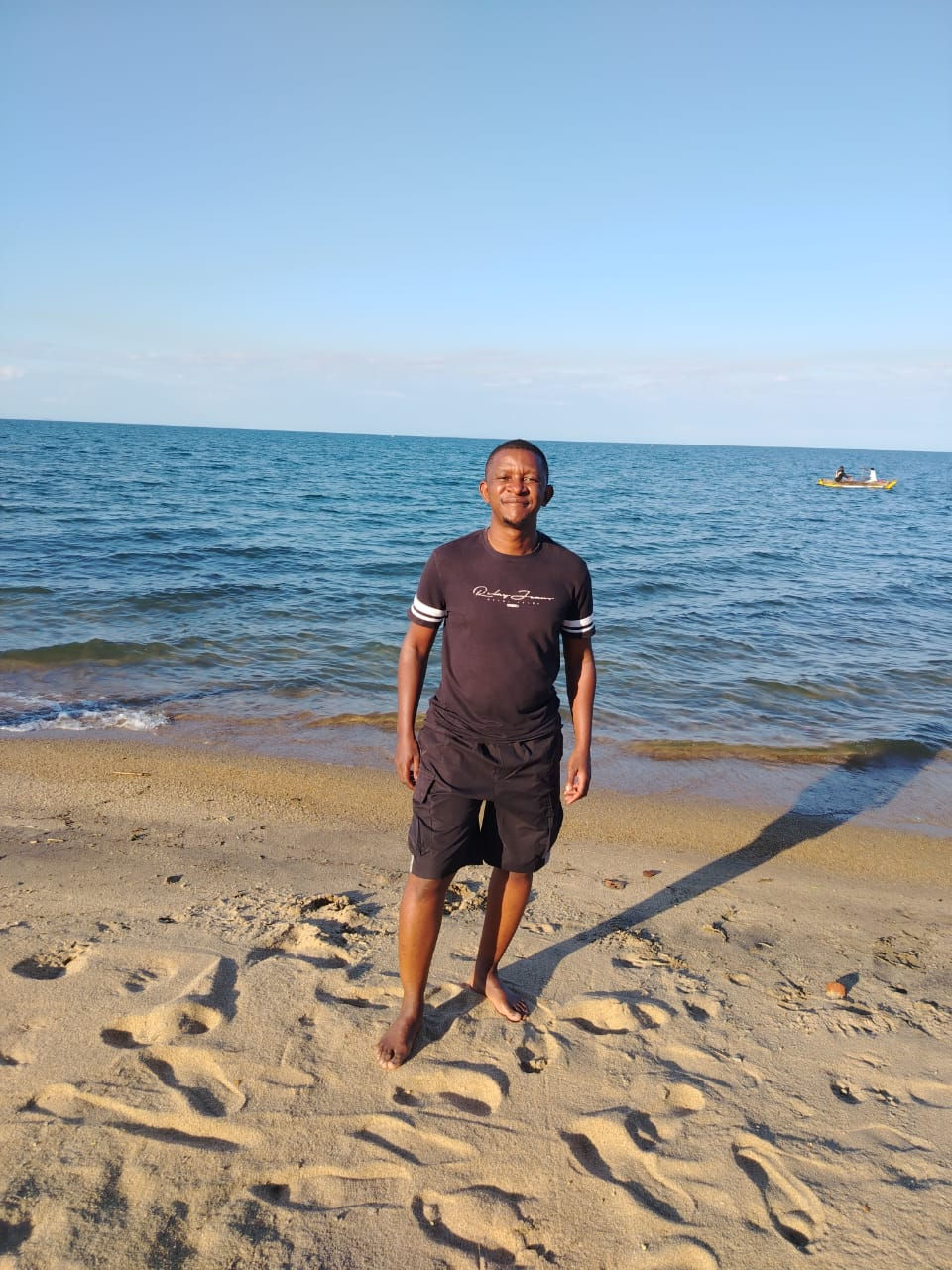 Man in black shirt and shorts stands smiling on sandy beach. Blue sea and sky in background with a small boat on the water.