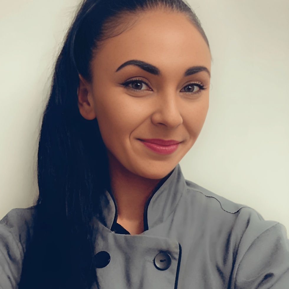 Bianca De Matos with long dark hair in a gray chef coat, set against a plain light background, exudes a confident, friendly demeanor.