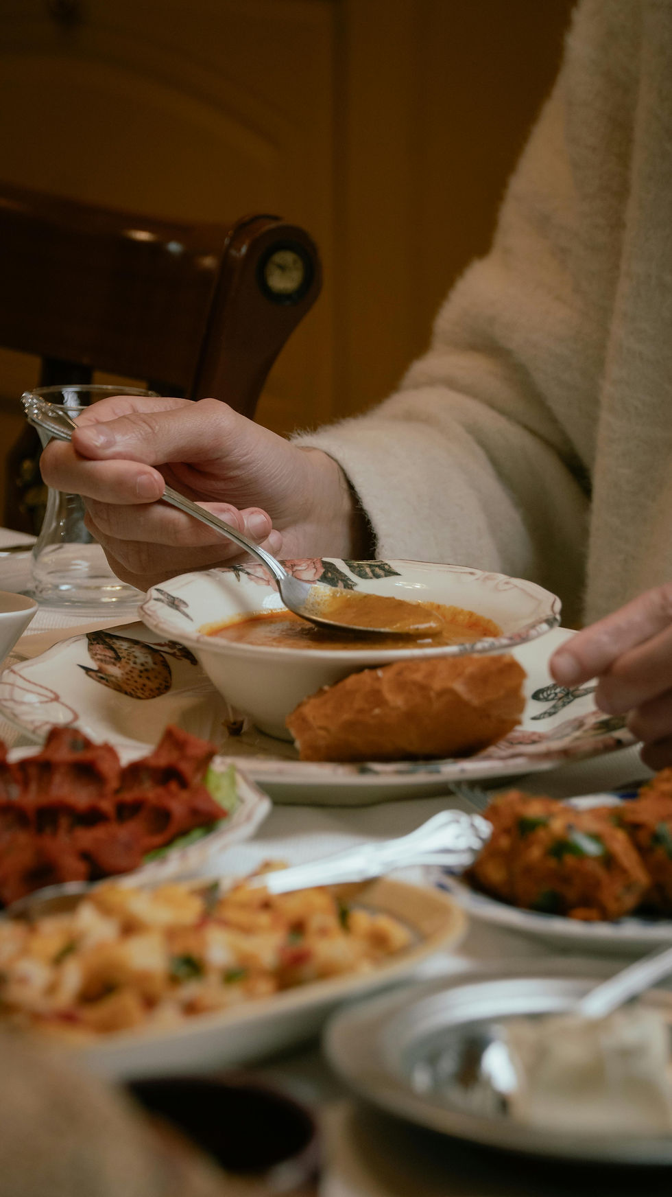 A person in a cozy sweater holds a spoon over a bowl of soup. Various dishes and bread are on the table, creating a warm dining atmosphere.