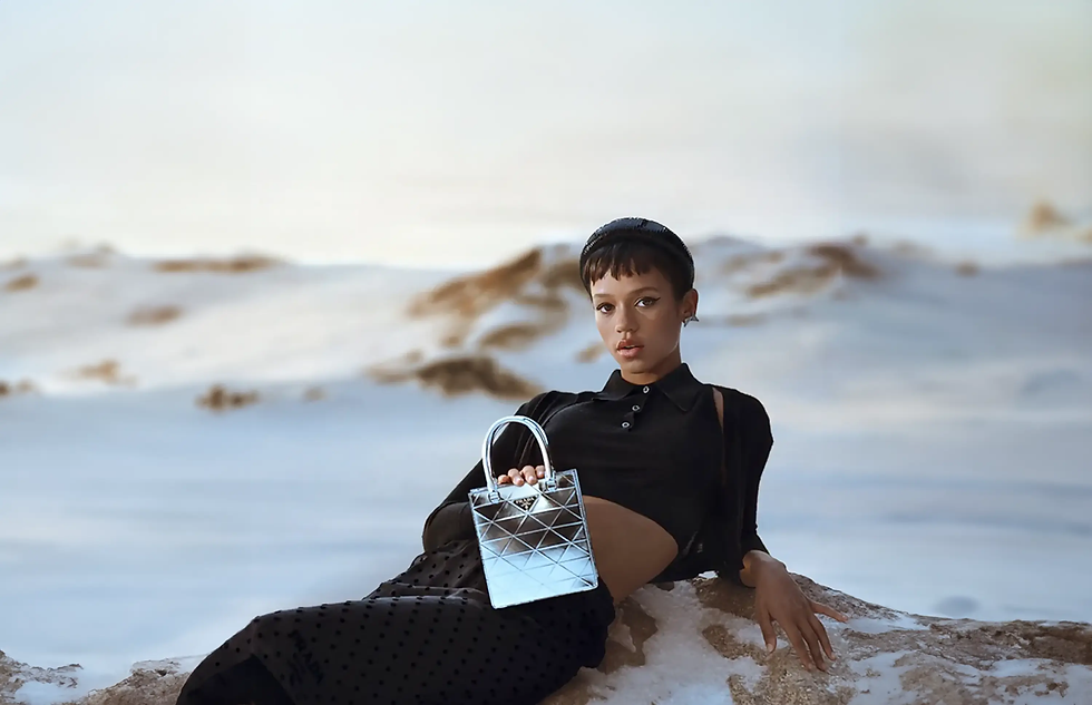 A fashion model reclining on sandy terrain in a desert-like landscape, wearing a black long-sleeve outfit and holding a small metallic handbag, with pale dunes and sky softly blurred in the background.