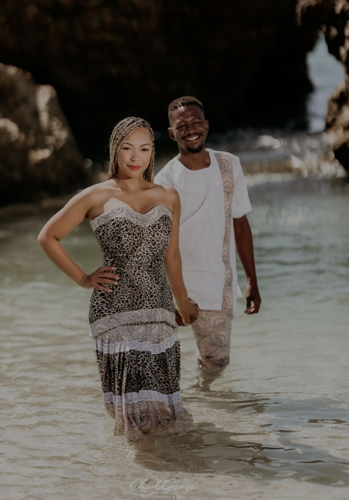 A smiling couple walks holding hands by the beach. The woman wears a leopard print dress; the man in a white outfit. Sunlit rocky background.