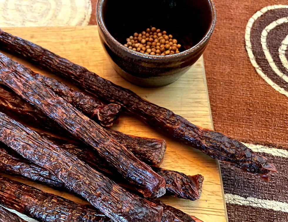 Dried sausages on a wooden board with a bowl of coriander seeds. Brown and beige patterned fabric in the background adds warmth.