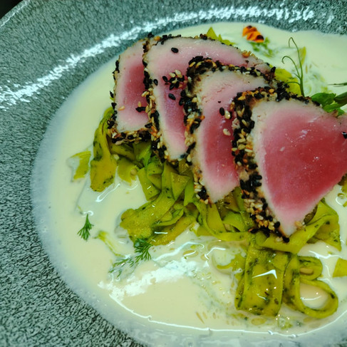Seared sesame-crusted tuna slices over green noodles in creamy white sauce on a speckled gray plate