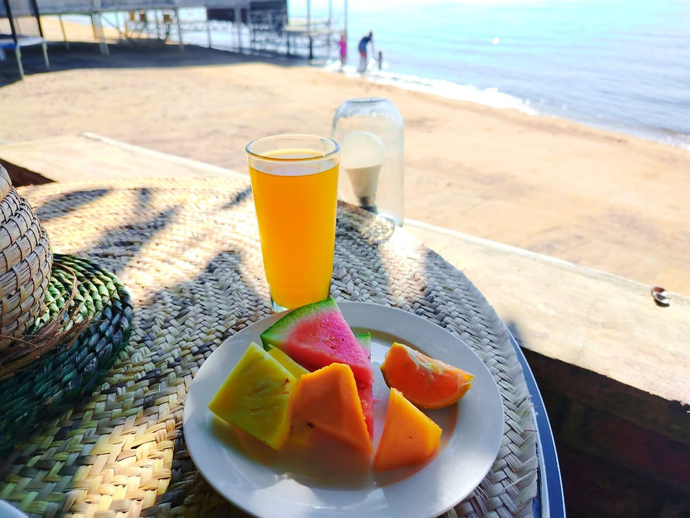 Plate of colorful fruit slices and a glass of orange juice on a woven mat, set on a beachside table with ocean in the background.