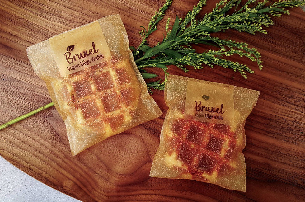 Two packaged vegan Liege waffles lie on a wooden surface beside green sprigs. The packaging reads "Bruxel." Warm, cozy ambiance.