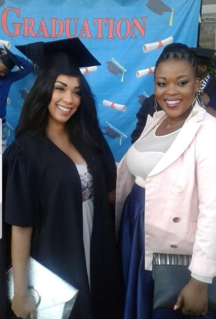 Two women smiling at a graduation event. One wears a cap and gown, holding a silver clutch. Backdrop reads "GRADUATION" with scrolls.