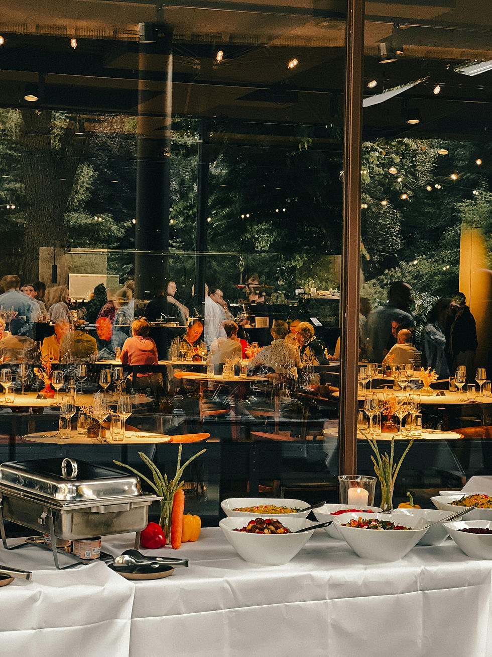 Elegant buffet table with salads and a chafing dish in a glass-walled restaurant, diners chatting under warm lights.
