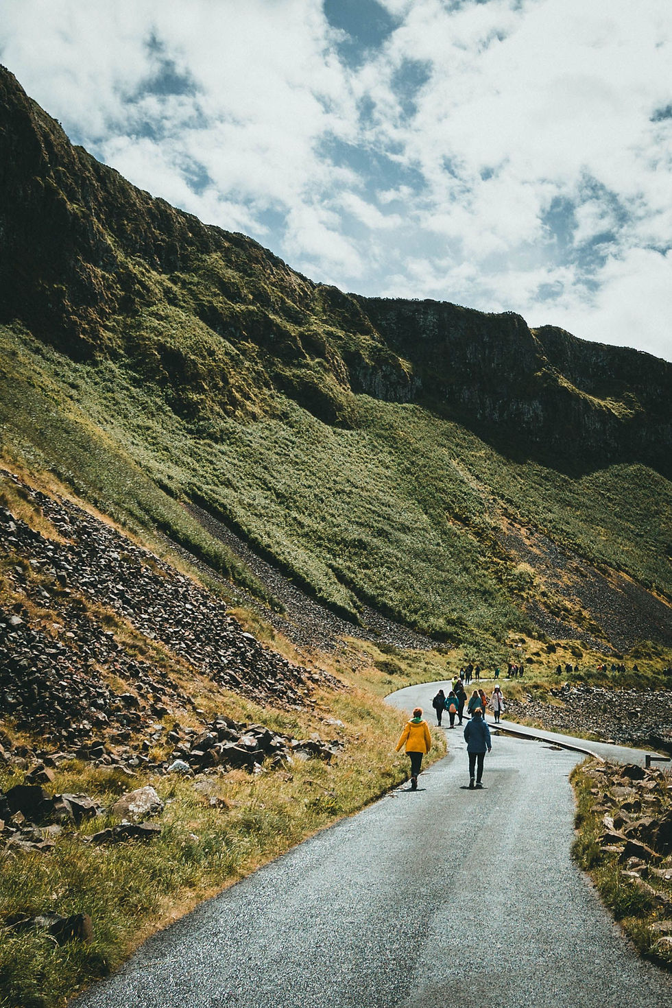 Hikers walk along a winding mountain road beside steep green cliffs under a cloudy sky.