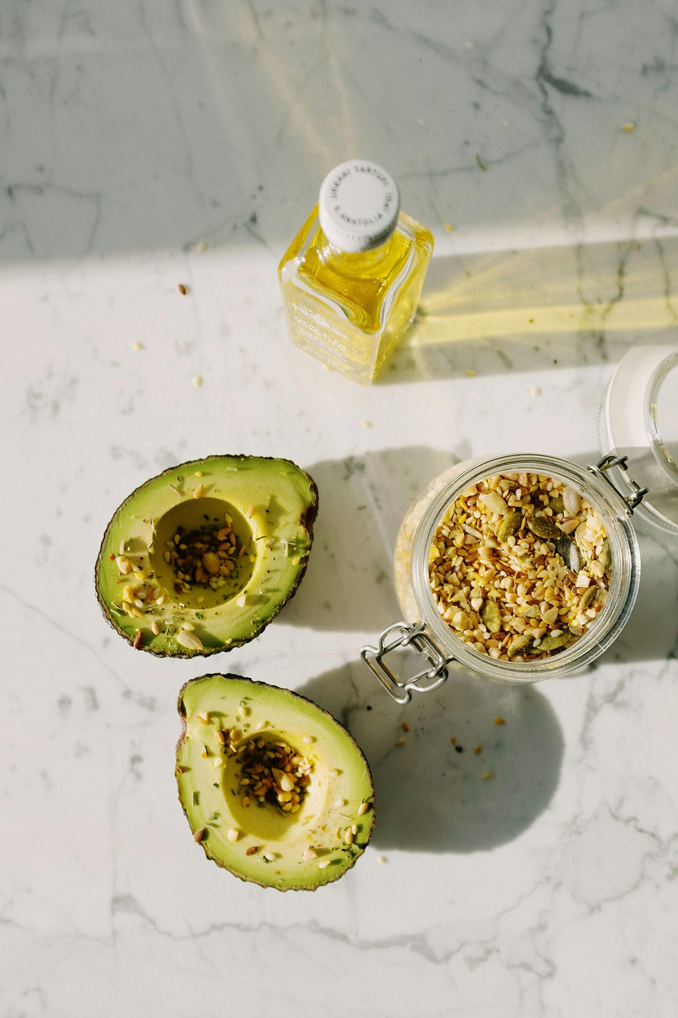 Halved avocado sprinkled with seeds, jar of mixed seeds, and a bottle of olive oil on a sunlit marble counter.