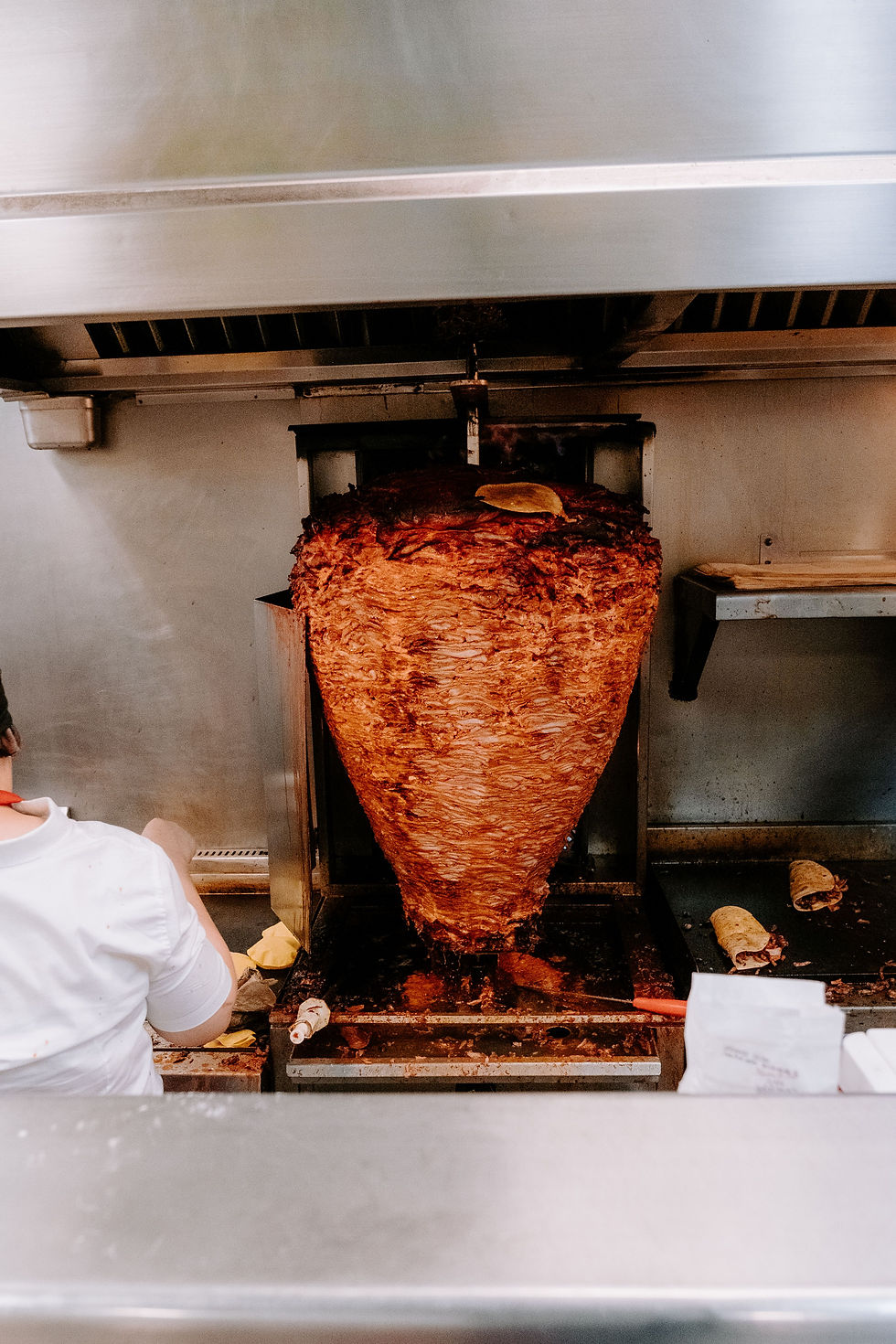 Cook in a kitchen stands by a huge vertical spit of roasted meat, with metal counters and trays around it.