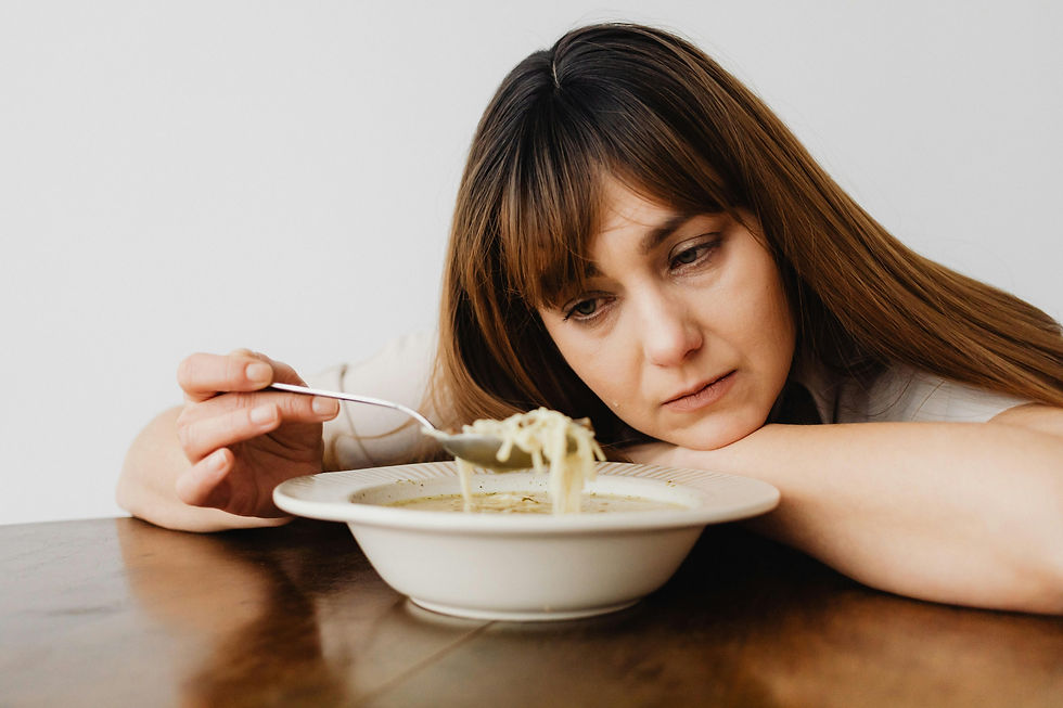 Woman resting head on table, looking at a bowl of noodles with a spoon. She appears pensive. Neutral background.