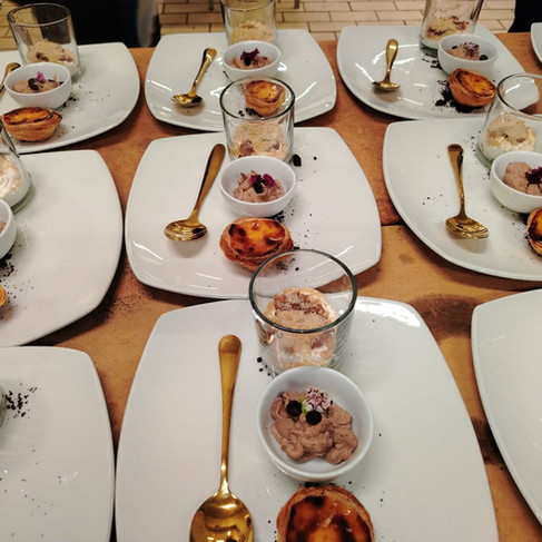 Rows of plated desserts with gold spoons and glass cups on a catering table, showing custard tarts and mousse.