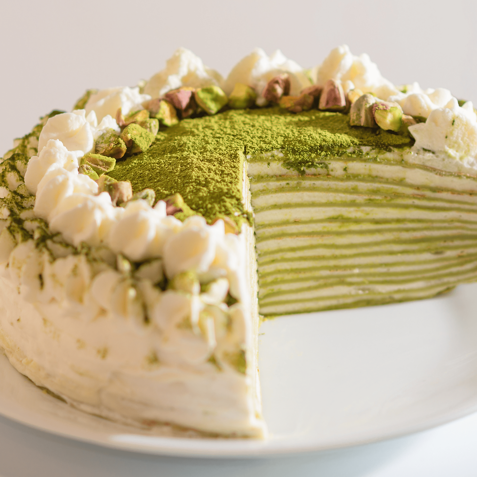 Matcha layered cake topped with whipped cream and pistachios, a slice removed. Green and white layers visible on a white plate.
