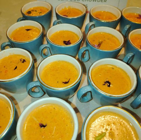Rows of blue cups filled with orange seafood chowder on a table, with a sign reading SEAFOOD CHOWDER in the background.
