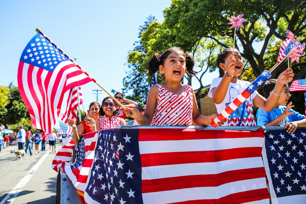 Children waving American flags and pinwheels on a parade float decorated with stars and stripes. They appear joyful under a sunny sky.