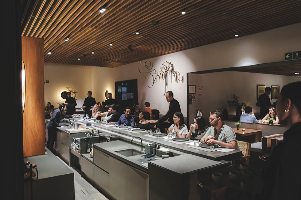 Dim upscale restaurant with diners seated around a long counter, waiters serving, under warm wood slat ceiling lighting