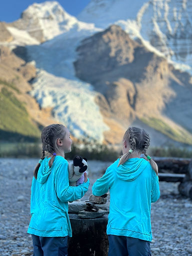 Two girls in turquoise jackets, one holding a plush toy, gaze at a mountain with snow. Rocky foreground, clear blue sky. Peaceful mood.