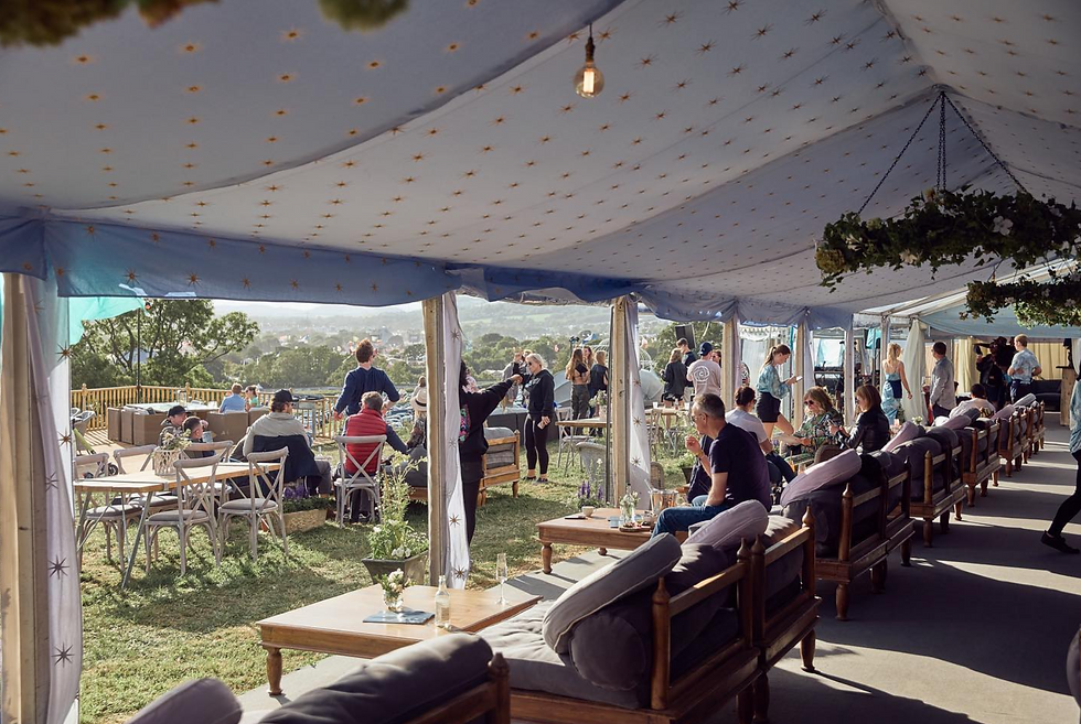 People relax under a star-patterned tent with sofas and tables, enjoying an outdoor event with a scenic view. Mood is lively and social.