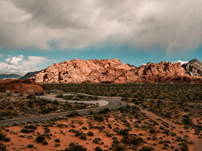 Desert landscape with a winding road leading to red rocky hills under a cloudy sky. Sparse vegetation dotting the orange terrain. Tranquil mood.