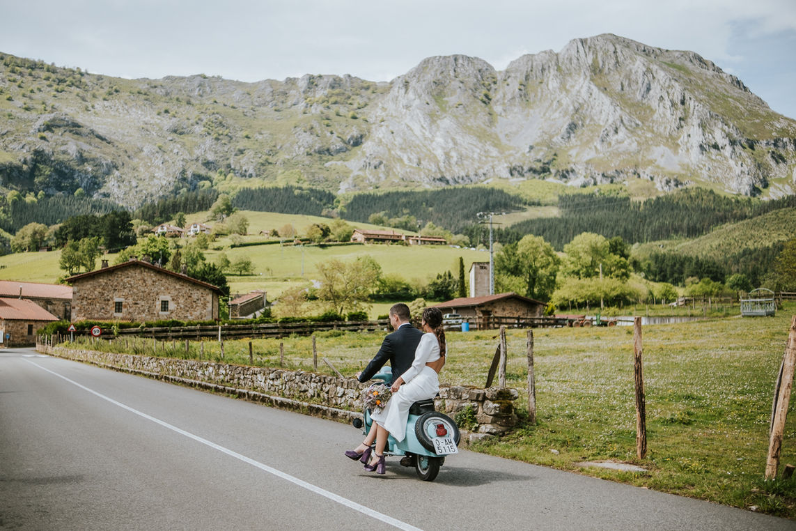 "Novios montados en una moto vintage recorriendo un paisaje rural con montañas de fondo durante una boda en Bizkaia."