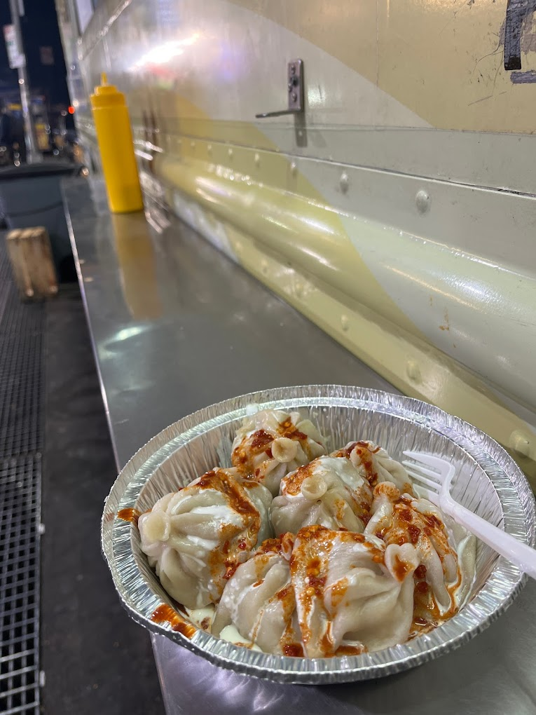 Tray of dumplings with spicy sauce sits on a metal counter beside a mustard bottle at night. Yellow and silver hues dominate the scene.