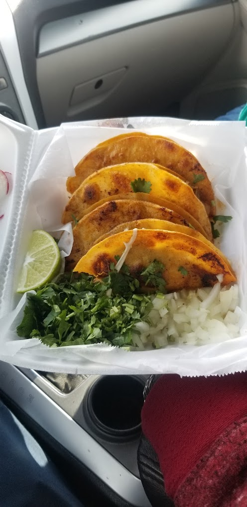 Four crispy tacos in a container with cilantro, onion, and lime. Set on a car seat; vibrant colors suggest a fresh, appetizing meal.