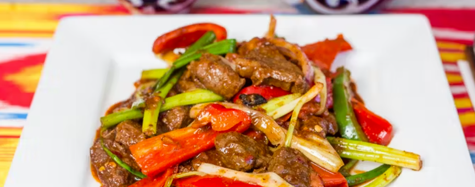 Stir-fried beef with red peppers, scallions, and onions on a white plate. Background has colorful striped patterns, creating a vibrant mood.
