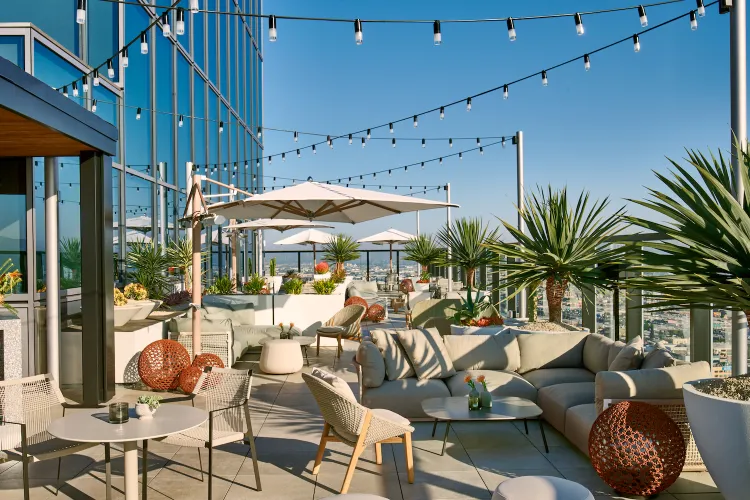 Rooftop patio with modern furniture, string lights, potted plants, and umbrellas. Clear blue sky enhances the relaxed, inviting atmosphere.