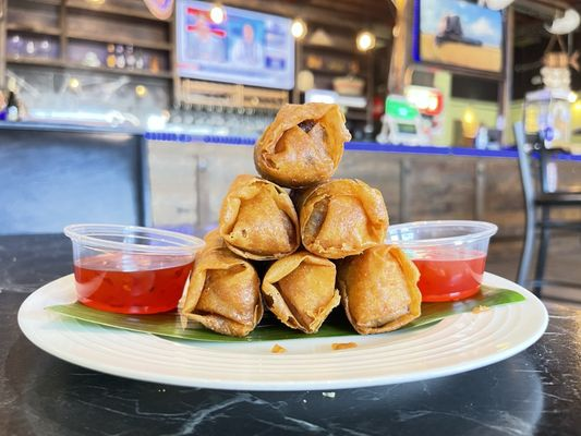 Spring rolls stacked on a plate with two cups of red sauce in a cozy restaurant. Bar in background with muted TV screens.