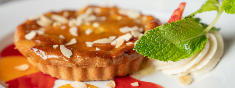 Tart with almond slices on a plate, garnished with cream, mint leaves, and red berry sauce. Bright and appetizing presentation.