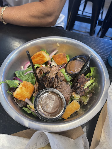 Salad in a metal bowl with grilled beef, croutons, and dressing containers. Arm in background, set on a dark table in a casual setting.