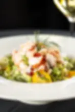 Lobster salad on a white plate with fresh greens, yellow tomatoes, and dill garnish. Background is dark; glass visible on the side.