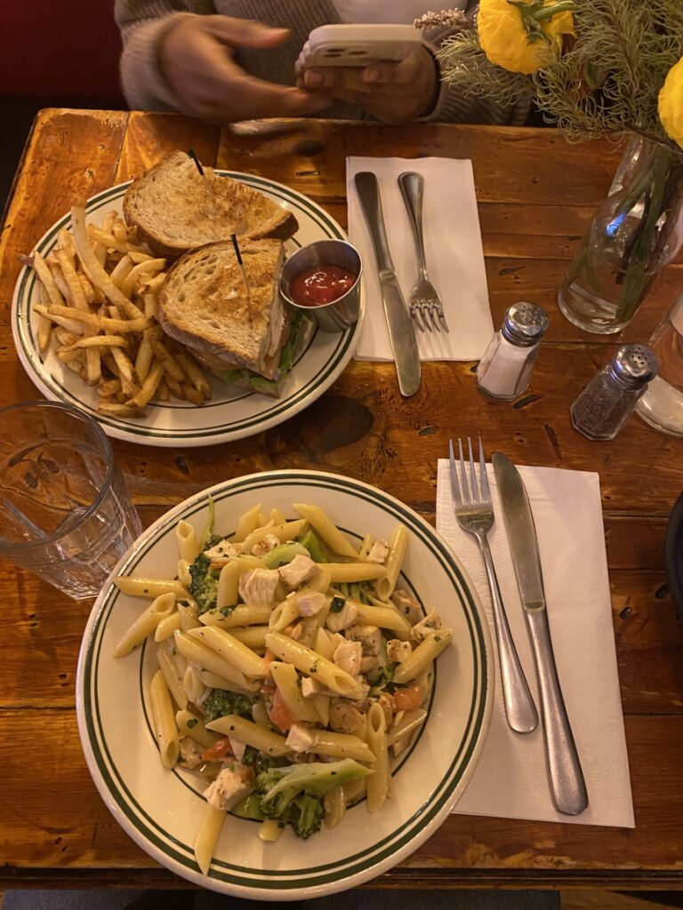 Pasta and sandwich with fries on a wooden table. A person is holding a phone. Yellow flowers in a vase. Casual dining setting.