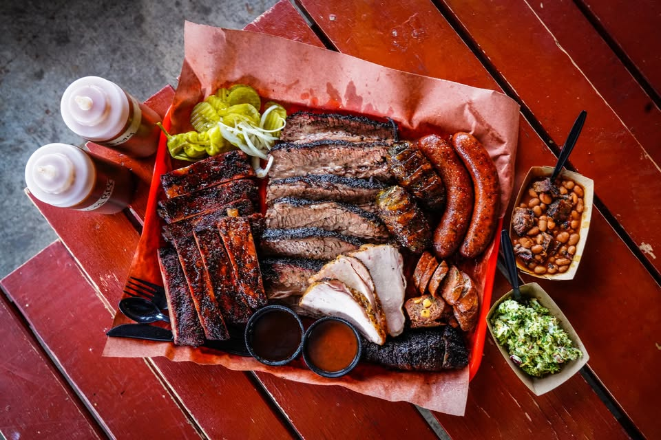 BBQ platter with ribs, brisket, sausage, pickles, coleslaw, and beans on a red tray. Two sauce bottles on a wooden table.