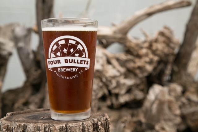 A pint glass of amber beer labeled "Four Bullets Brewery, Richardson, TX" on a tree stump with a blurred wood background. Rustic mood.