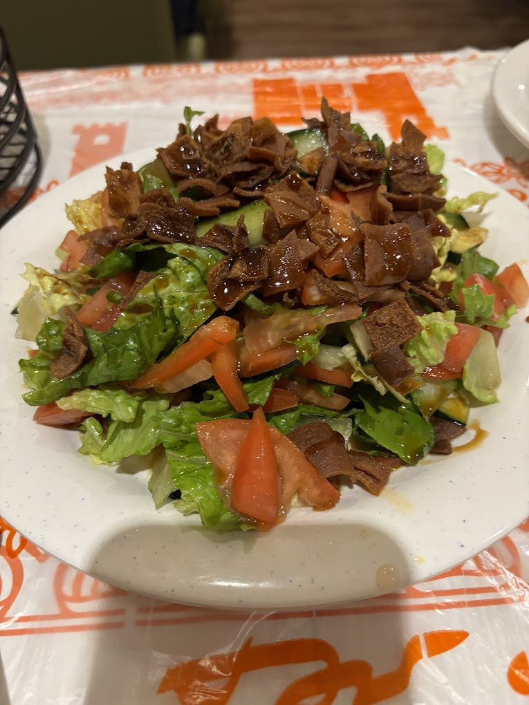 Colorful salad with lettuce, tomato, cucumber, and crispy pieces drizzled with dressing on a white plate. Orange and white tablecloth.
