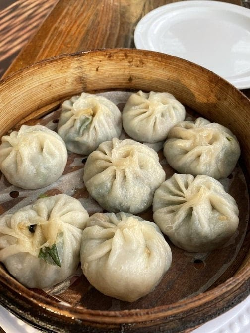 Steamed dumplings in a bamboo basket on a wooden table, with a white plate in the background. Warm, inviting dining setting.
